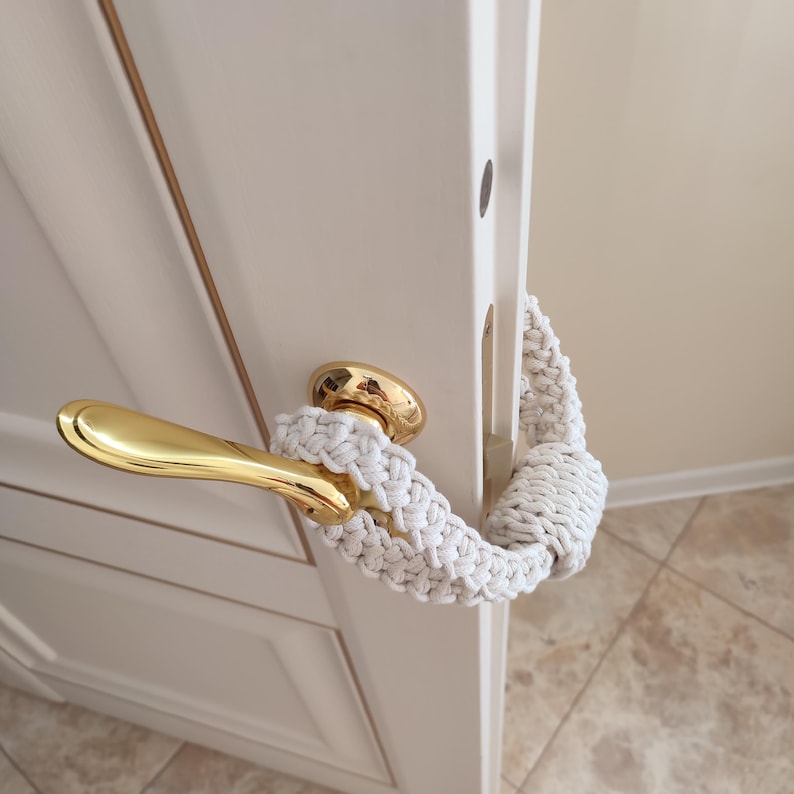 May include: A white macrame door handle cover featuring a braided cord design with a knotted end. The accessory is wrapped around a polished gold-tone metal lever handle attached to a white door, providing a textured grip and decorative accent.