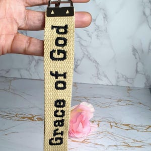Könnte beinhalten: Beigefarbener Stoff-Schlüsselanhänger mit dem in Schwarz gestickten Schriftzug "Grace of God". Er hat einen Metallverschluss und einen Ring. Eine rosa Rose ist im Hintergrund. Ein religiöses Accessoire.