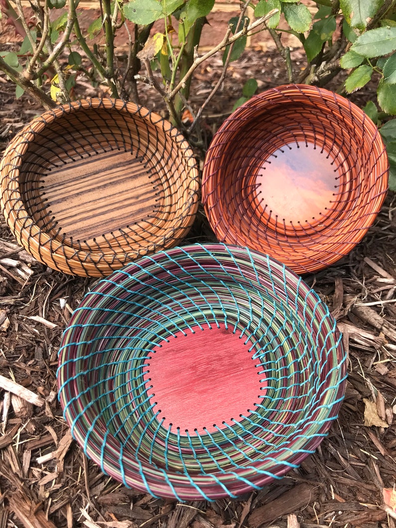 Puede incluir: Tres cestas tejidas con diferentes tejidos de colores y patrones. Las cestas est&aacute;n hechas de materiales naturales y tienen una forma redonda. Las cestas est&aacute;n sentadas en un lecho de mantillo marr&oacute;n.