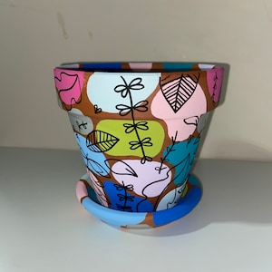 May include: A hand-painted terracotta planter with a brown base and a colourful design featuring blue, green, pink, and yellow abstract shapes and leaves. The planter has a matching saucer.