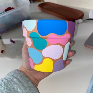 May include: A terracotta flower pot with a colourful abstract design. The pot is painted with various shades of blue, green, pink, yellow, and purple.