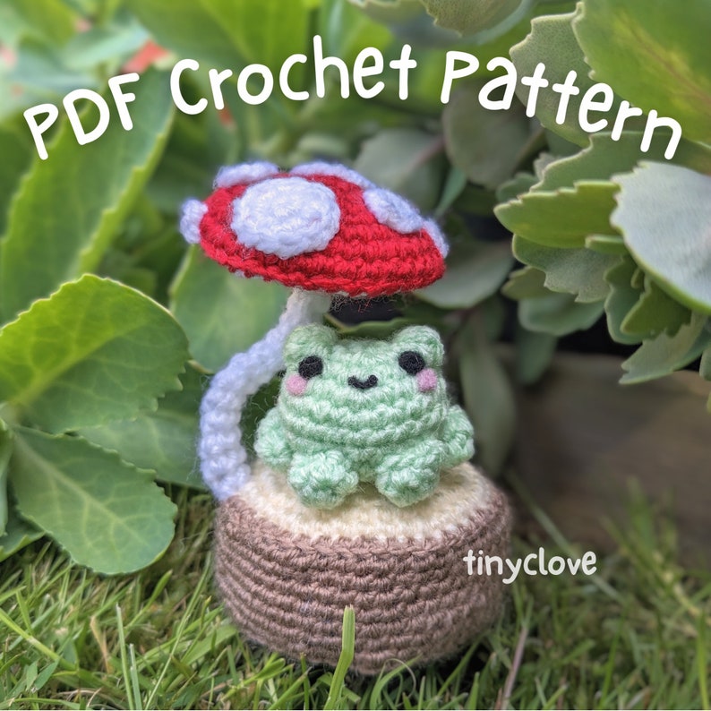 May include: Crochet pattern for a green frog sitting under a red and white mushroom with white spots. The mushroom and frog are sitting on a brown and beige crocheted log. The text "tinyclove" is visible in the lower right corner of the image.