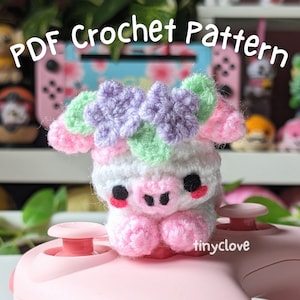 May include: A crocheted pig amigurumi wearing a flower crown with purple and green flowers. The pig is sitting on a pink surface with the text "tinyclove" visible.