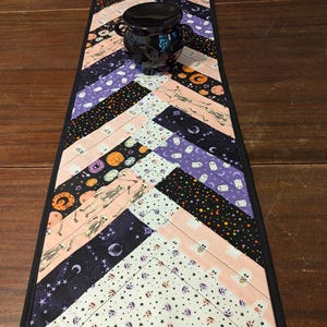 May include: A Halloween-themed table runner with a black border and a chevron pattern. The fabric features various Halloween motifs, including skeletons, ghosts, pumpkins, and stars, in shades of black, purple, pink, and white. A black cauldron sits on top.