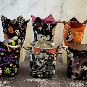 May include: Six Halloween-themed fabric cup cozies in various colors and patterns. The cozies are made of fabric with different designs, including skulls, ghosts, bats, and pumpkins.