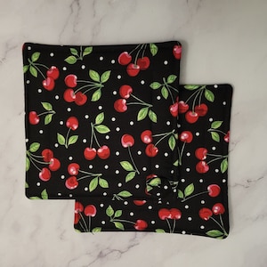 Könnte beinhalten: Zwei schwarze quadratische Topflappen mit einem weißen Tupfenmuster und roten Kirschen mit grünen Blättern.
