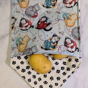 May include: A light blue fabric potato storage bag with a cat and yarn print. The bag has a white paw print bottom and contains two potatoes. The cats are in various colors and poses.