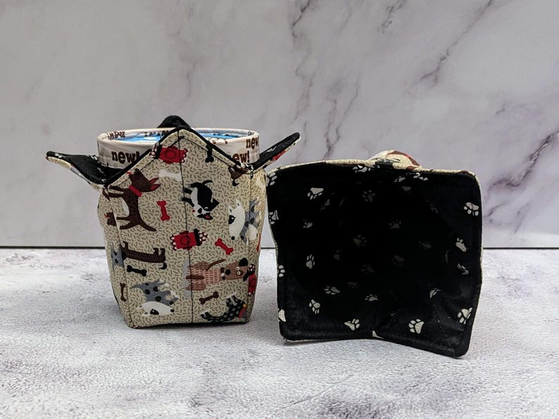 May include: Two fabric bowl cozies. One is tan with dog and bone patterns, holding a mug with "new!" text. The other is black with white paw prints. These are designed to protect hands from hot bowls.