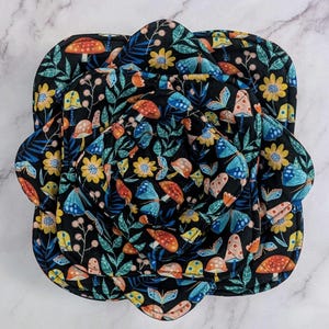 Może przedstawiać: Zestaw czterech czarnych podkładek pod garnki z kwiatowym wzorem i fantazyjnym designem. Wzór zawiera kolorowe grzyby, motyle i kwiaty w odcieniach żółci, niebieskiego, pomarańczowego i zielonego. Podkładki są ułożone w konfiguracji zagnieżdżonej.