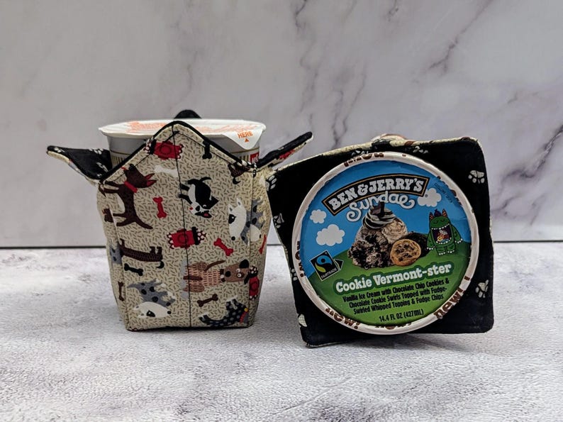 May include: Two fabric cozies for ice cream containers. One features a dog pattern on a beige background, the other is black with paw prints. A Ben & Jerry's ice cream container is visible.
