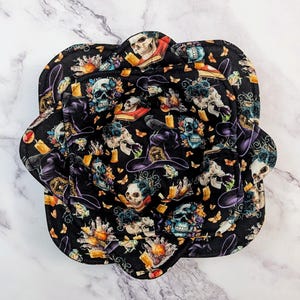 May include: Black and purple fabric bowl with a witchy skull and hat design. The bowl is lined with a black fabric and has a scalloped edge.