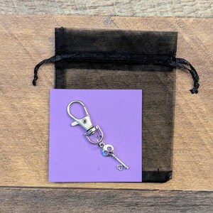 May include: A silver-toned metal keychain featuring a lobster clasp and a small key charm with a skull-shaped bow and a heart-shaped bit, displayed on a square purple card inside a black sheer drawstring pouch.