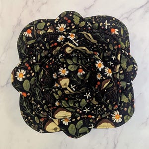 Może przedstawiać: Zestaw czarnych akcesoriów kuchennych z kwiatowym wzorem, w tym podkładka pod garnek i osłona na miskę. Wzór przedstawia białe stokrotki, grzyby i zielone liście. Przedmioty znajdują się na białej marmurowej powierzchni.