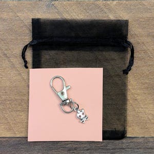 May include: A silver-toned metal keychain featuring a small skull and crossbones charm with a bow detail, attached to a lobster claw clasp. The keychain is displayed on a square light pink card placed in front of a black sheer drawstring pouch.