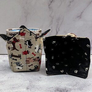 May include: Two fabric bowl cozies. One is tan with dog and bone patterns, holding a mug with "new!" text. The other is black with white paw prints. These are designed to protect hands from hot bowls.