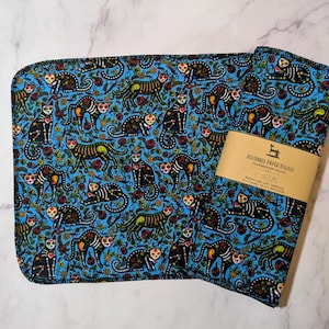 May include: A set of reusable paper towels featuring a bright blue background with a repeating pattern of black cats decorated with colorful floral and skeletal motifs. A tan paper band is wrapped around the towels with text reading Reusable Paper Towels, 10 x 10 inches, Set of 6, 100% Cotton.
