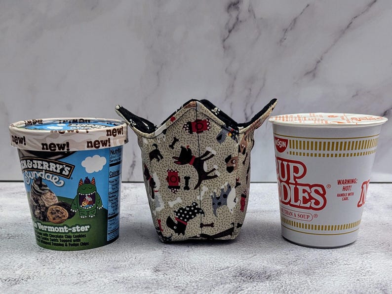 May include: An image featuring a Ben & Jerry's ice cream container, a fabric bowl with a dog pattern, and a Nissin Cup Noodles container. The ice cream container has a blue and white label. The fabric bowl has a dog and bone pattern. The Cup Noodles container is white with red text.