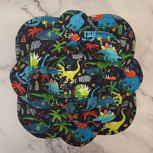 May include: A set of three black fabric placemats with a dinosaur print. The placemats feature a variety of dinosaurs in shades of blue, green, yellow, and red on a black background. The placemats are shaped like a flower with rounded edges.