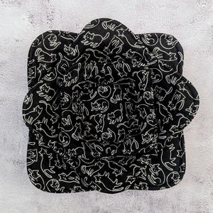 Può includere: Presa pentola in tessuto nero con un motivo di gatto bianco. La presa pentola ha la forma di un fiore con più petali arrotondati.