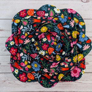 May include: A dark green, floral-patterned pot holder in the shape of a flower. The fabric features colorful illustrations of mushrooms, butterflies, and flowers in shades of red, yellow, pink, and blue. The pot holder is on a light wood surface.