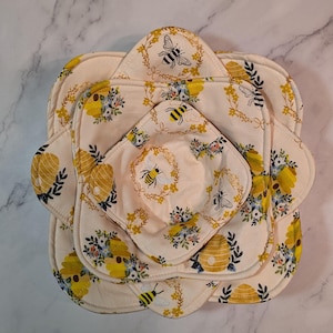 Może przedstawiać: Zestaw czterech kremowych, materiałowych osłon na miski z falistymi krawędziami i nadrukiem pszczół i kwiatów. Osłony ułożone są w zagnieżdżony wzór, prezentujący różne rozmiary i wzory. Ilustracje pszczół są żółte i czarne.