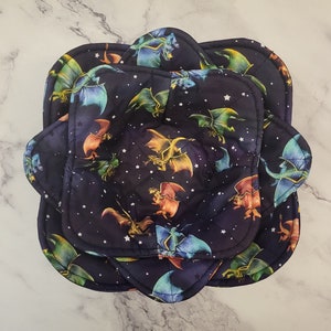 Pode incluir: Conjunto de quatro pegadores acolchoados com fundo azul escuro e um padrão repetido de dragões coloridos e estrelas brancas. Os pegadores têm a forma de uma flor com bordas arredondadas.
