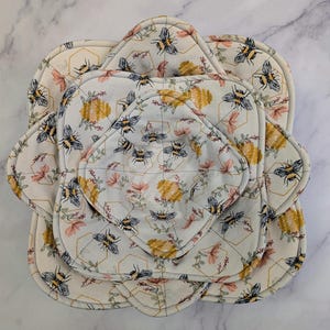 Może przedstawiać: Zestaw czterech pikowanych podkładek z tkaniny z wzorem pszczół i plastra miodu. Podkładki są białe z powtarzającym się wzorem pszczół, plastrów miodu i elementów kwiatowych w kolorze żółtym, różowym i szarym. Różne rozmiary.