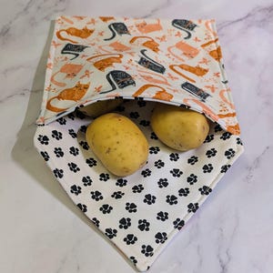 May include: A reusable, white fabric produce bag with a cat pattern on the top half and black paw prints on the bottom half. Two potatoes are visible inside the bag. The cats are orange, black, and pink.