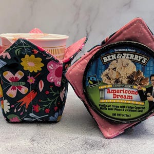 Op de afbeelding: Twee stoffen komhoezen, een met een kopje noedels en de andere met een pint (473ml) Ben & Jerry's Americone Dream ijs. De hoezen hebben bloempatronen en roze accenten.