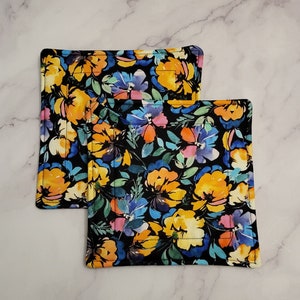 Könnte beinhalten: Zwei schwarze Topflappen aus Stoff mit einem floralen Muster in Gelb-, Blau- und Rosatönen. Das Muster ist im Aquarellstil auf schwarzem Hintergrund gehalten.