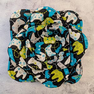 May include: Three black fabric bowl cozies with a blue and green pattern of video game controllers. The cozies are layered on top of each other.