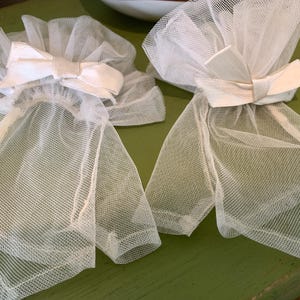 May include: Two white tulle hair accessories with a bow detail. The accessories are made of a sheer, net-like fabric and are arranged on a green surface. The bows are made of a solid white fabric.