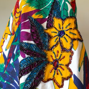 May include: A white fabric shirt with a tropical print featuring large yellow, green, and purple leaves. The shirt has a beaded flower applique on the sleeve with yellow, red, and blue beads.