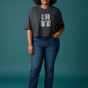May include: A charcoal gray cropped t-shirt featuring the white text DJ SPIN THAT SHIT printed on the front, paired with dark blue denim jeans and white low-top canvas sneakers. The shirt has short sleeves and a crew neckline.
