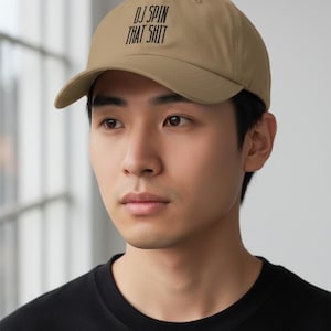 May include: A tan baseball cap featuring the black text DJ SPIN THAT SHIT embroidered on the front panel. The cap has a structured crown, a curved brim, and a button on the top center. The design is centered on the front of the hat.