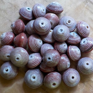 Op de afbeelding: Een verzameling handgemaakte kralen met een spiraalvormig ontwerp. De kralen zijn voornamelijk rood en wit, met een cilindrische vorm en een gat in het midden. De kralen liggen op een houten oppervlak.