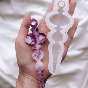 Puede incluir: Adorno de resina morada y blanca hecho a mano con detalles de purpurina, junto a su molde de silicona. El adorno tiene un diseño abstracto único con un lazo en la parte superior para colgar. El molde es blanco y tiene una forma similar.
