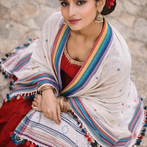 Può includere: Una donna indossa un abito rosso e uno scialle color crema con strisce arcobaleno e nappe. Indossa orecchini d'oro, un copricapo e braccialetti. Un fiore rosso adorna i suoi capelli. Lo scialle ha dettagli blu, verdi e rossi.