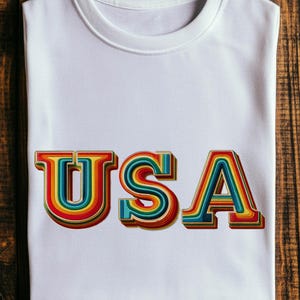 Pode incluir: Camiseta branca com a palavra "USA" em um design retrô. As letras são multicoloridas, com camadas de cores do arco-íris e um efeito 3D. A camiseta está em uma superfície de madeira.