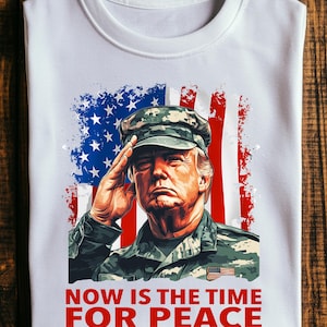 May include: White t-shirt featuring a graphic of a man saluting in military attire, set against a backdrop of the American flag. The text "NOW IS THE TIME FOR PEACE" is printed in red at the bottom of the shirt.