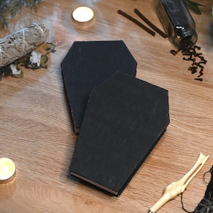 May include: Two black coffin-shaped boxes with flat lids, positioned one slightly overlapping the other. The boxes feature a matte black finish and a distinct hexagonal silhouette, showcasing a simple, functional design for storage or display purposes.