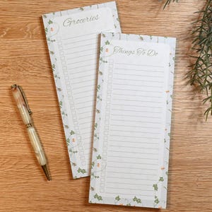 May include: Two rectangular notepads with floral borders and lined pages. One notepad reads "Groceries," the other "Things To Do." A pen with a cream and pink marbled barrel rests on a wooden surface.