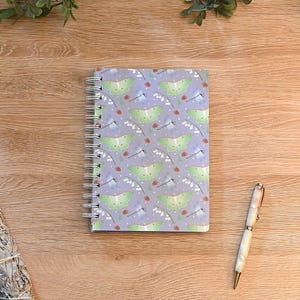 May include: A spiral-bound notebook with a lavender cover featuring a repeating pattern of green moths, white flowers, and red berries. A white and gold pen rests on a wooden surface, along with a bundle of sage.