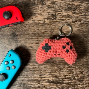May include: A crocheted pink video game controller keychain with black buttons. The keychain is attached to a silver key ring.