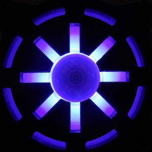 May include: A black circular object with a purple light emanating from the center. The light shines through eight spokes, creating a starburst pattern.