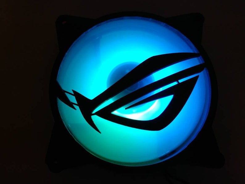 May include: A black computer fan with a blue and green LED light. The fan has a black logo with the letters "ROG" on it.