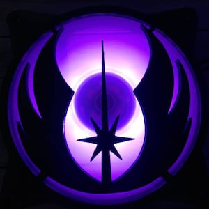 May include: A black computer fan with a purple glowing design. The design features a star and a stylized emblem, illuminated by a bright purple light. The fan blades are visible within the central circle.
