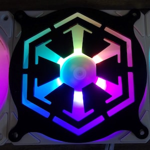 May include: A black computer fan with a white and rainbow LED light design. The fan has a cutout in the shape of the Star Wars Imperial symbol.