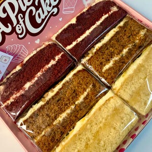 Op de afbeelding: Een roze doos met het opschrift A Piece of Cake bevat zes rechthoekige plakken cake, elk afzonderlijk verpakt in doorzichtig plastic. Het assortiment bestaat uit twee red velvet plakken, twee worteltaartplakken en twee lichte biscuitplakken.