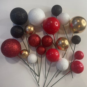 Könnte beinhalten: Eine Auswahl an dekorativen Kugel-Picks in verschiedenen Größen und Farben. Die Kugeln umfassen glitzerndes Rot und Schwarz, einfarbiges Rot, Weiß und Gold. Jede Kugel ist an einem dünnen grünen Drahtstiel befestigt, geeignet für Blumengestecke oder Bastelarbeiten.